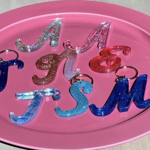 May include: A collection of resin alphabet letter keychains in various colors including blue, red, and copper, featuring glitter and iridescent finishes. The letters include A, J, n, &, T, S, and M, some with attached silver-toned metal key rings.