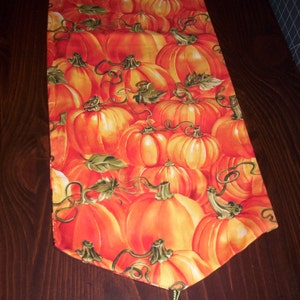 May include: A decorative table runner featuring a repeating pattern of orange pumpkins with green stems and leaves. The runner has a tassel at the end and is set on a dark brown wooden surface.
