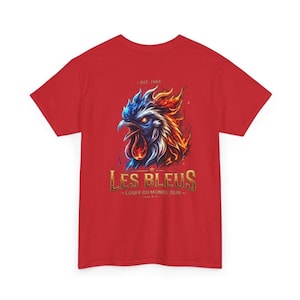 May include: Red t-shirt featuring a stylized rooster design with blue, orange, and white flames. The text "LES BLEUS - COUPE DU MONDE 2026" is displayed in gold lettering. The shirt also has the text "- EST. 1904 -" above the rooster.