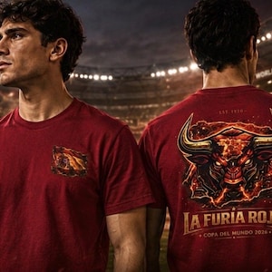 May include: Two red t-shirts. One features a Spanish flag emblem, the other a bull graphic with "LA FURIA ROJA" and "COPA DEL MUNDO 2026" text. The shirts are worn by a person in a stadium setting.