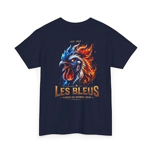 May include: Navy blue t-shirt featuring a fiery rooster graphic with blue and orange flames. The text "LES BLEUS" is displayed in gold, with "COUPE DU MONDE 2026" below. The shirt also includes the text "- EST. 1904 -".