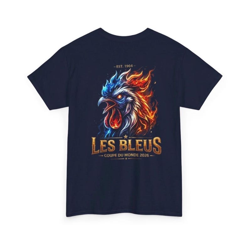 May include: Navy blue t-shirt featuring a fiery rooster graphic with blue and orange flames. The text "LES BLEUS" is displayed in gold, with "COUPE DU MONDE 2026" below. The shirt also includes the text "- EST. 1904 -".