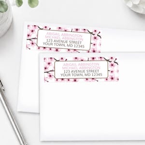 May include: Two white envelopes with address labels featuring a floral design of pink cherry blossoms. The labels include the text "ABIGAIL ABBINGTON, MICHAEL ABBINGTON, 123 AVENUE STREET, YOUR TOWN, MD 12345". A silver pen rests on the left.