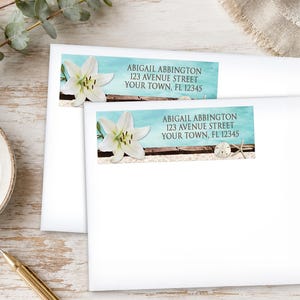 May include: Two white envelopes with address labels featuring a lily flower, sand, and a light blue background. The labels read "ABIGAIL ABBINGTON 123 AVENUE STREET YOUR TOWN, FL 12345". One label has a starfish and a sand dollar.