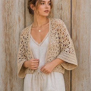 Könnte beinhalten: Beige Häkeljacke mit offenem Front- und Dreiviertelärmel. Die Jacke wird über einem weißen Camisole und einem beigen Overall getragen. Das Outfit wird mit einer goldenen Halskette ergänzt. Der Hintergrund ist eine hellbraune Holzwand.