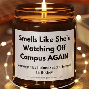 May include: A brown glass jar candle with a white label featuring the black text: Smells Like She's Watching Off Campus AGAIN. Warning: May Induce Sudden Interest In Hockey. The candle sits on a round wooden coaster with a lit wick.