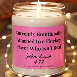 May include: A clear glass candle jar with a pink label featuring the text: Currently Emotionally Attached to a Hockey Player Who Isn't Real, John Logan, #22. The candle sits on a round wooden base and contains white wax.