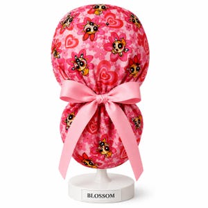 May include: A pink scrub cap featuring a repeating pattern of cartoon characters, pink cherry blossoms, and red hearts. The fabric cap has a gathered back secured with a light pink satin ribbon tied into a bow. The cap is displayed on a white stand.
