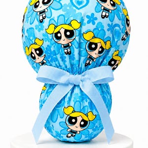 May include: A light blue scrub cap featuring a repeating pattern of the character Bubbles from The Powerpuff Girls, accented with blue flowers and hearts. The cap includes a gathered back with a light blue ribbon tied in a bow at the center.
