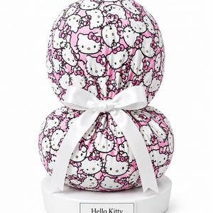 May include: A rounded, two-tiered fabric object featuring a repeating pattern of Hello Kitty faces on a pink background. The two sections are cinched together by a white satin ribbon tied in a bow. The object rests on a white circular base with a rectangular plaque displaying the text Hello Kitty.