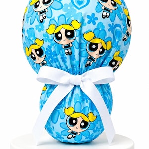 May include: A light blue scrub cap featuring a repeating pattern of the character Bubbles from The Powerpuff Girls with yellow pigtails and blue outfits, accented by blue flowers and hearts, finished with a white ribbon tied in a bow at the back.