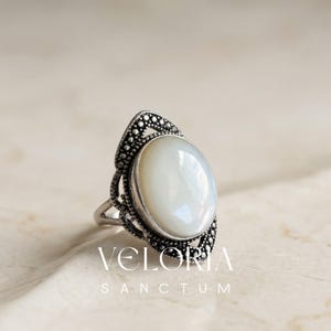 Può includere: Un anello color argento con una pietra bianca iridescente di forma ovale, incastonata in una cornice metallica decorativa con intricati dettagli a perline. La fascia è divisa e il metallo ha una finitura ossidata. Il testo VELORIA SANCTUM è visibile.