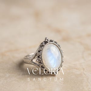 May include: A silver-toned ring featuring an oval-shaped, iridescent moonstone cabochon set within a decorative beaded metal border. The band has a split design, and the text VELORIA SANCTUM is printed in white letters across the lower portion.