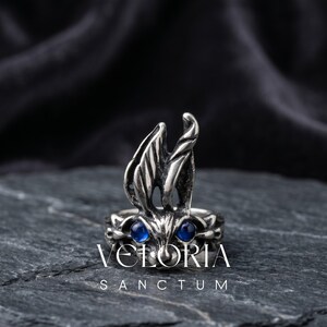 Puede incluir: Un anillo de metal plateado con una cabeza de conejo esculpida, orejas largas y erguidas y dos ojos redondos de piedra preciosa azul oscuro. La superficie del metal tiene un acabado oscuro y oxidado. El texto VELORIA SANCTUM aparece debajo.