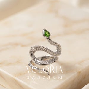 May include: A silver-toned snake ring featuring a textured, scale-like pattern along the body and a single pear-shaped green gemstone set in the head. The text VELORIA SANCTUM is visible in white lettering at the base of the coiled snake design.