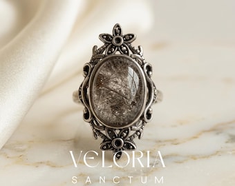 May include: An oval-shaped silver ring with a clear stone containing black inclusions. The ring has an ornate floral design around the stone and on the shoulders. The text "VELORIA SANCTUM" is printed below the ring.