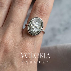 May include: A silver ring featuring an oval-shaped mother-of-pearl inlay with a carved floral design of tulips. The centrepiece is surrounded by a delicate beaded border. The text VELORIA SANCTUM is printed in white at the bottom of the image.