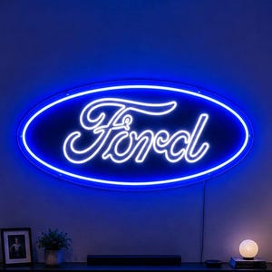 Peut inclure: Une grande enseigne néon Ford de forme ovale fixée au mur, émettant une lueur bleue vive. L'enseigne présente le logo emblématique Ford en écriture blanche, entouré d'une bordure néon bleue continue, créant un affichage lumineux éclatant.