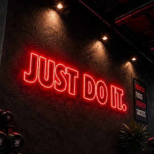以下が含まれることがあります： 「JUST DO IT.」と書かれた赤いネオンサインがあるジムの内観。サインは暗い質感の壁に取り付けられています。ダンベルが前景に見え、床は濃い赤色です。