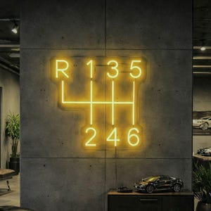 May include: A neon sign in the shape of a car's manual transmission gear shift, with the numbers R, 1, 3, 5, 2, 4, and 6 illuminated in a bright yellow color. The sign is mounted on a gray concrete wall.