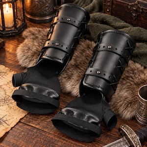 May include: A pair of black fingerless gauntlets featuring structured forearm guards with adjustable straps, metal studs, and crisscross lacing. The hand portion is made of a flexible fabric with a thumb hole and a wide elastic band across the palm.