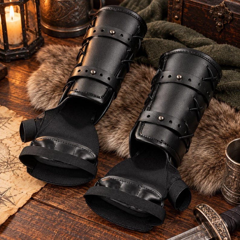May include: A pair of black fingerless gauntlets featuring structured forearm guards with adjustable straps, metal studs, and crisscross lacing. The hand portion is made of a flexible fabric with a thumb hole and a wide elastic band across the palm.