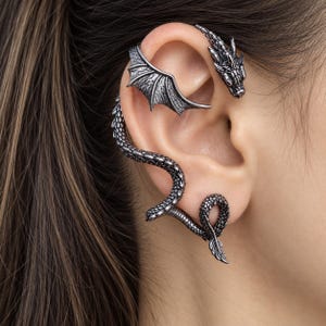 Può includere: Un ear cuff color argento a forma di drago, caratterizzato da una testa di drago dettagliata nella parte superiore, un'ala testurizzata che si estende sul cartilagine e una lunga coda squamata che si curva lungo il bordo esterno del lobo.