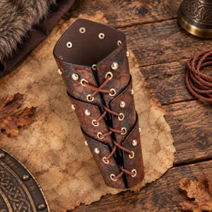 Puede incluir: Un brazalete de cuero marrón con superficie texturizada, ojales de metal dorado y cordones marrones que se entrelazan verticalmente por el centro. El protector de brazo cilíndrico está asegurado con remaches metálicos en los bordes.