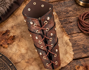 May include: A brown leather bracer featuring a textured surface, gold-toned metal eyelets, and brown cord lacing that runs vertically down the center. The cylindrical arm guard is secured with multiple metal rivets along the edges and sides.