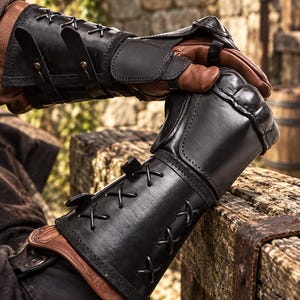 May include: A pair of black leather gauntlets featuring brown leather inner gloves, black leather forearm guards with crisscross lacing, and adjustable buckle straps. The gauntlets have reinforced knuckle protection and a structured design.