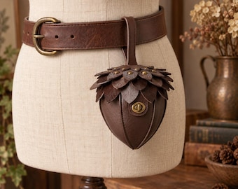 May include: A dark brown leather belt featuring an attached pouch shaped like an acorn. The pouch has a textured cap with layered leaf-like details, a smooth rounded base, and a brass-toned metal twist-lock closure. The belt has a matching buckle.
