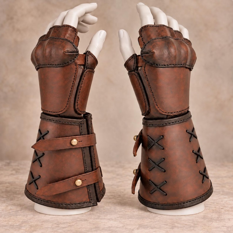May include: A pair of brown fingerless gauntlets featuring a reinforced knuckle plate, a structured forearm section with black crisscross lacing, and two horizontal leather straps secured with small brass-colored metal studs on the outer side.