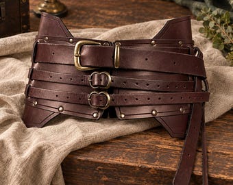 May include: A wide dark brown leather corset belt featuring three horizontal straps with antique brass buckles and metal rivets. The structured design includes multiple adjustment holes along each strap and a long, tapered end piece.