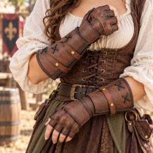 May include: A pair of fingerless brown leather gauntlets featuring dark brown lacing, adjustable straps with gold-toned metal rivets, and a structured forearm design. The gauntlets are worn over a white long-sleeved shirt and a brown corset with lacing.