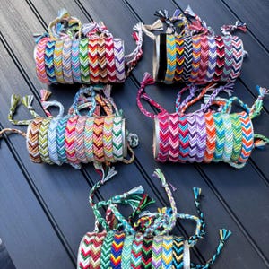 Peut inclure: Cinq rouleaux cylindriques en carton contenant chacun une collection de bracelets d'amitié tissés. Les bracelets présentent des motifs en chevron dans diverses combinaisons de couleurs incluant des fils rouges, bleus, verts, jaunes, roses, violets, noirs et blancs.