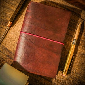 May include: A handmade, reddish-brown leather journal with a pink elastic closure. A brass pen and a leather working tool are next to the journal on a wooden surface. The journal is closed and ready to be written in.