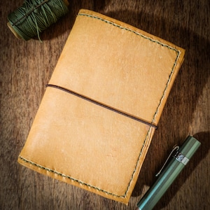 May include: A tan leather notebook cover featuring visible green stitching along the edges and a thin dark brown elastic band wrapped horizontally around the center. A spool of green thread and a metallic green fountain pen are positioned nearby.