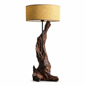 May include: A table lamp featuring a dark brown, gnarled wooden base with a natural, textured surface. A thin black metal rod extends upward from the center of the wood to support a cylindrical, mustard yellow fabric lampshade with a woven pattern.