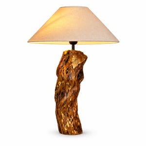 May include: A table lamp featuring a natural, textured wooden log base with a polished brown finish and a wide, conical beige fabric lampshade. The lamp emits a warm glow from beneath the shade, highlighting the organic wood grain.