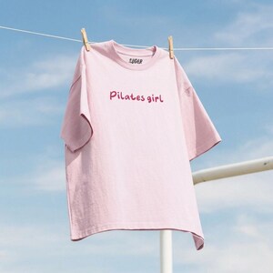 May include: A light pink t-shirt with the text "Pilates girl" in a cursive font. The word "SUGAR" is printed above the neckline. The shirt is hanging on a clothesline with wooden clothespins against a blue sky.
