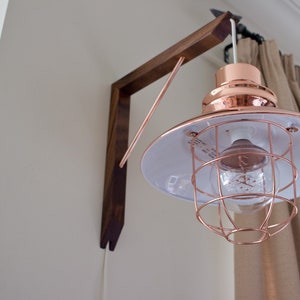 Puede incluir: Una l&aacute;mpara colgante de tono cobre con una pantalla blanca y una jaula de alambre. La luz est&aacute; montada en un soporte de pared de madera.