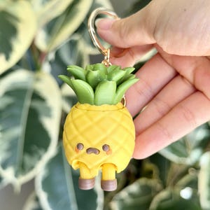 May include: A yellow pineapple-shaped keychain with a green leafy top. The pineapple has a sad facial expression with small brown eyes and a downturned mouth, along with two small yellow legs wearing brown boots attached to the bottom.