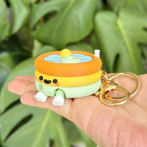May include: A three-tiered inflatable pool keychain featuring an orange top ring, a yellow middle ring, and a light green base. The pool has a smiling face, a small yellow floating ball, and two white feet extending from the bottom, attached to a gold ring.