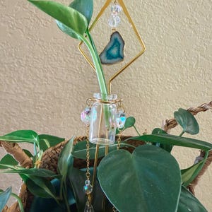 Könnte beinhalten: Eine klare Glas-Blumen-Vase mit einem goldfarbenen Metallrahmen und dekorativen Akzenten. Der Rahmen hält eine türkis-weiße Geodenscheibe und klare Kristallperlen. Die Vase enthält einen grünen Pflanzenstiel mit großen grünen Blättern.
