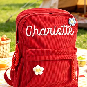 Può includere: Uno zaino rosso in velluto a coste con il nome "Charlotte" ricamato in bianco. Lo zaino ha una tasca frontale, una maniglia superiore e due toppe floreali. La borsa è su una coperta da picnic.