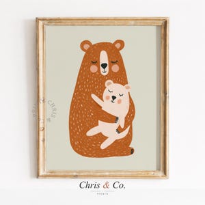Può includere: Una stampa artistica incorniciata raffigurante un'orsa che abbraccia un cucciolo d'orso. Gli orsi sono marroni e color crema, con gli occhi chiusi e le guance rosee. La stampa ha una cornice dorata e il testo "Chris & Co." in basso.