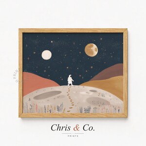 Puede incluir: Una impresión artística enmarcada representa a un astronauta caminando sobre una superficie lunar bajo un cielo estrellado. La obra de arte tiene un marco de madera marrón claro, dos lunas y el texto "Chris & Co."