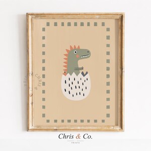 May include: A framed print featuring a cartoon dinosaur hatching from an egg. The dinosaur is green with a red crest, and the egg is white with black spots. The background is tan, and the frame is gold. The text "Chris & Co." is at the bottom.