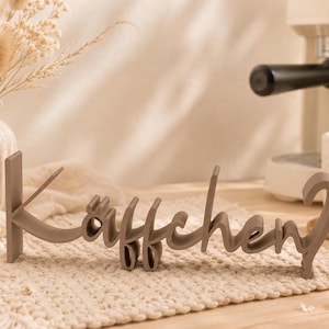 Könnte beinhalten: Ein taupefarbenes, dreidimensionales freistehendes Schild mit dem deutschen Wort "Käffchen?" in einer Schreibschrift. Die Buchstaben sind miteinander verbunden und haben eine glatte, matte Oberfläche, die zum Aufstellen gedacht ist.