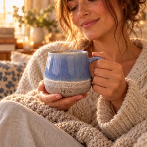 Può includere: Una tazza in ceramica con una sezione superiore blu lucida e una base grigia testurizzata e maculata. La tazza presenta un manico curvo e il nome Olivia stampato con un semplice carattere scuro sulla parte inferiore grigia esterna.